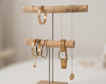 May include: A wooden jewelry display stand with two horizontal bars and a square base. The stand holds various gold-toned jewelry, including necklaces, bracelets, and a watch. The jewelry stand is made of light-colored wood and has a minimalist design.