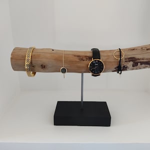 May include: A wooden jewelry stand with a natural bark finish. The stand features a long, branch-like piece of wood with a black metal base. The stand is decorated with a gold watch, a gold bracelet, a gold necklace, and a gold ring.