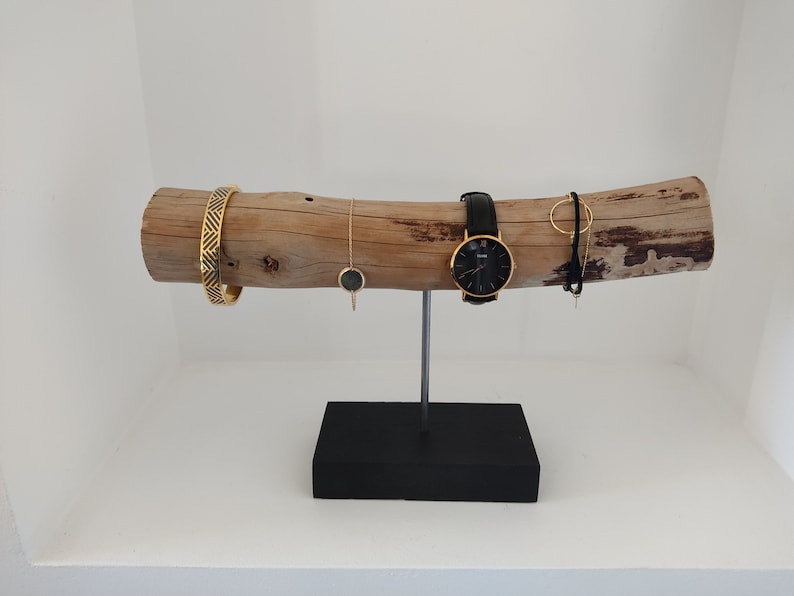 May include: A wooden jewelry stand with a natural bark finish. The stand features a long, branch-like piece of wood with a black metal base. The stand is decorated with a gold watch, a gold bracelet, a gold necklace, and a gold ring.