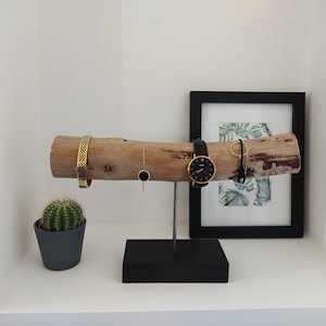 May include: A wooden jewelry stand with a natural bark finish. The stand features a long, horizontal branch with a black metal base. The branch is decorated with a gold bracelet, a gold necklace, a black watch, and a gold ring. A small potted cactus sits on a white shelf below the stand.