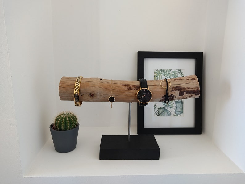 May include: A wooden jewelry stand with a natural bark finish. The stand features a long, horizontal branch with a black metal base. The branch is decorated with a gold bracelet, a gold necklace, a black watch, and a gold ring. A small potted cactus sits on a white shelf below the stand.