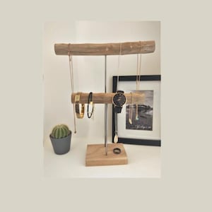 Peut inclure: Un présentoir à bijoux à deux niveaux composé de deux barres en bois flotté horizontales soutenues par une tige métallique centrale sur une base carrée en bois. Il porte des colliers dorés, une montre à cadran noir et un bracelet.