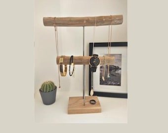 May include: A two-tiered jewelry stand featuring two horizontal driftwood bars supported by a central metal rod on a square wooden base. The stand holds several gold-toned necklaces, a black-faced wristwatch, and a bracelet, with a ring on the base.