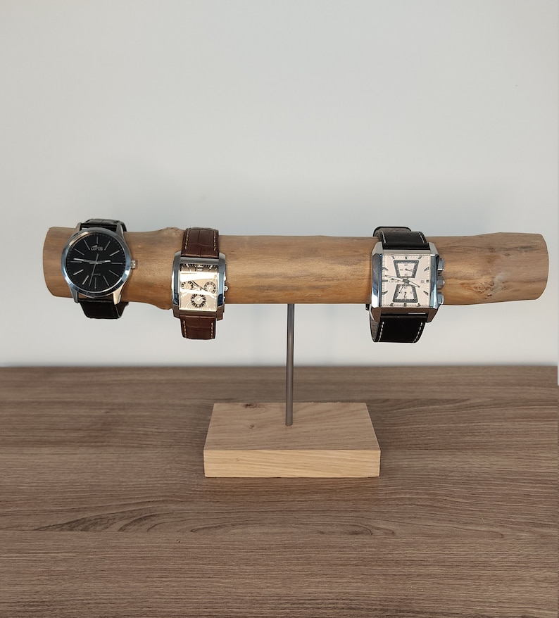 May include: A wooden watch stand with three watches on it. The stand is made of a natural branch of wood with a square wooden base. The watches are all different styles and colors.