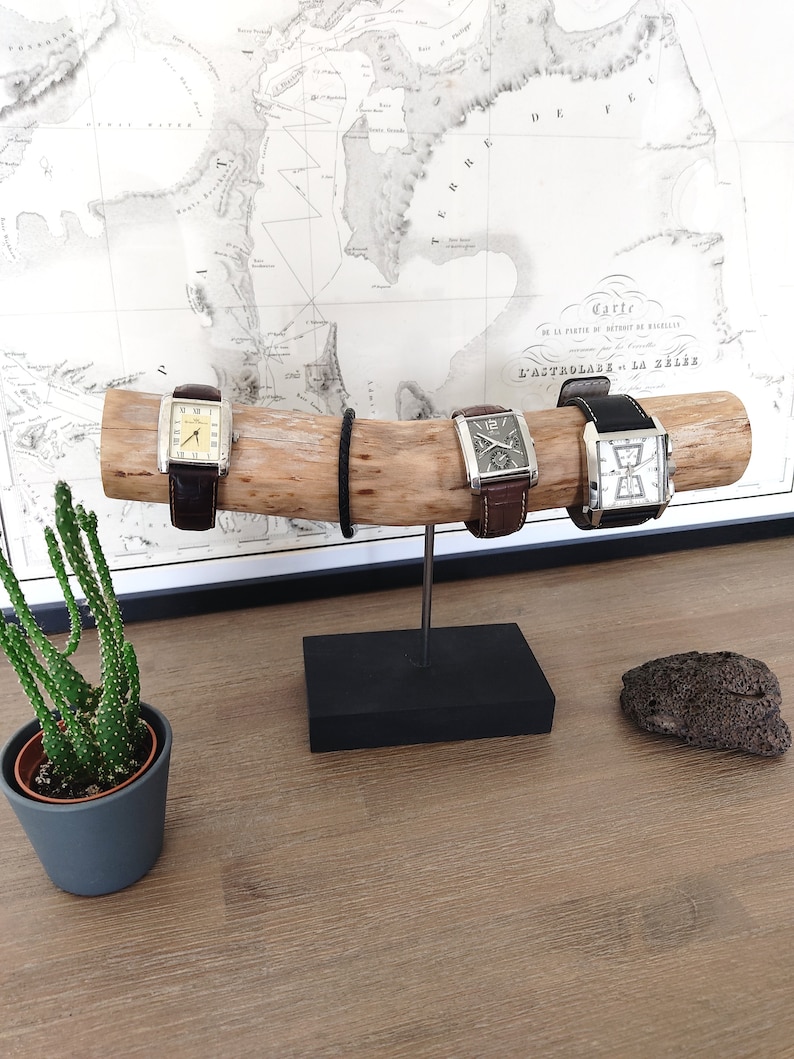May include: Three wristwatches with brown leather bands are displayed on a wooden stand. The stand is on a black base and is on a wooden surface. The watches have silver cases and white faces.