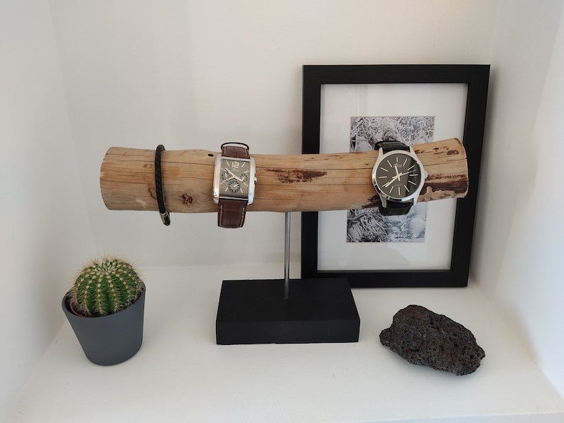 May include: A wooden watch stand with two watches, one with a brown leather band and the other with a black leather band. The stand is on a black base and there is a small cactus in a gray pot to the left.