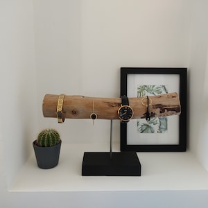 May include: A wooden jewelry stand with a black base. The stand features a natural branch with a gold bracelet, a gold necklace, a watch, and a gold ring. The stand is displayed on a white shelf with a small cactus plant in a gray pot.