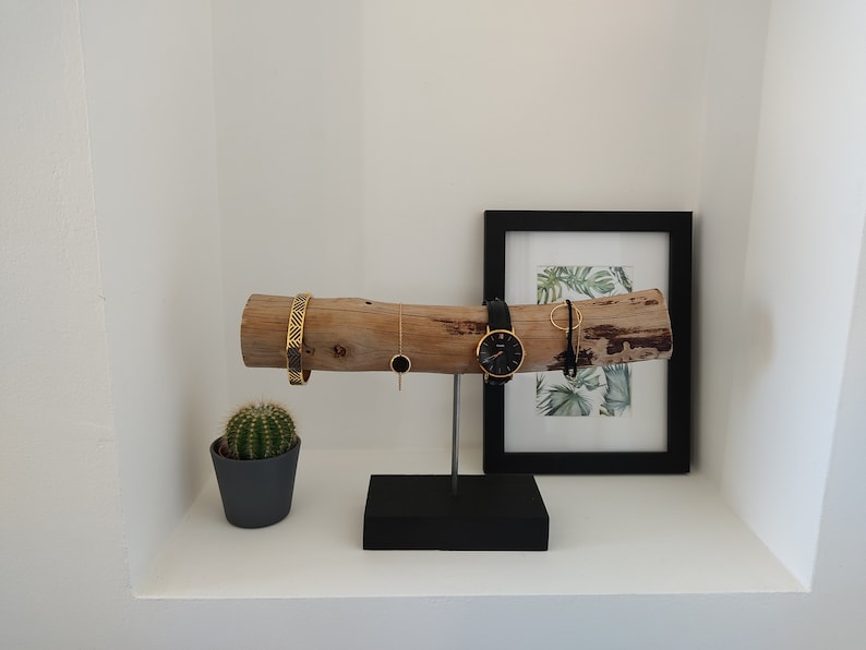 May include: A wooden jewelry stand with a black base. The stand features a natural branch with a gold bracelet, a gold necklace, a watch, and a gold ring. The stand is displayed on a white shelf with a small cactus plant in a gray pot.