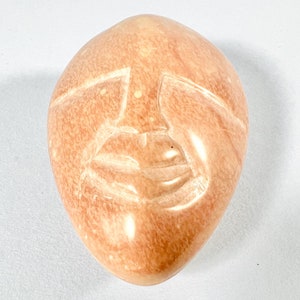 May include: A hand-carved stone sculpture of a face with a closed-mouth smile. The sculpture is made of a light brown stone with a smooth, polished finish.
