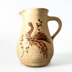 May include: A beige ceramic pitcher with a brown bird design. The pitcher has a rounded body and a handle.