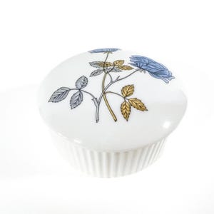 Puede incluir: Caja de porcelana blanca para alhajas con un diseño floral. La tapa presenta una rosa azul y dorada con hojas verdes.