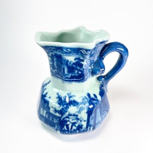 May include: A blue and white ceramic pitcher with a handle. The pitcher features a detailed landscape design in blue against a white background. The rim and interior are a light green color. The handle is a solid blue.