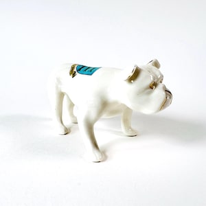 May include: A white ceramic bulldog figurine with gold accents on the ears and snout. A turquoise and gold design is on the dog's back. The bulldog is standing on all fours.