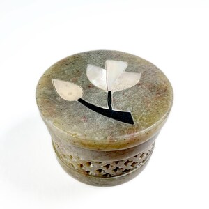 Op de afbeelding: Een ronde, olijfgroene stenen doos met een gebeeldhouwd bloemmotief rond de basis. Het deksel heeft een bloemontwerp ingelegd met witte en zwarte materialen. De doos is een decoratief opbergitem.