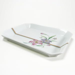 Pode incluir: Bandeja retangular branca com um design floral. A bandeja apresenta um arranjo floral rosa e roxo com folhas verdes e uma fina borda dourada e verde-azulado. A bandeja tem uma borda ligeiramente elevada.