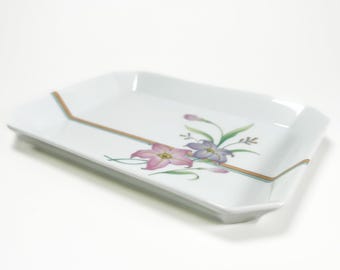 May include: White rectangular serving tray with a floral design. The tray features a pink and purple flower arrangement with green leaves and a thin gold and teal border. The tray has a slightly raised edge.