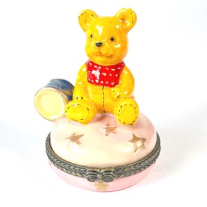 Puede incluir: Una figura de oso amarillo con una bufanda roja se sienta sobre una caja de baratijas rosa decorada con estrellas. La caja tiene un borde metálico decorativo y un cubo azul y amarillo. Un artículo decorativo encantador.