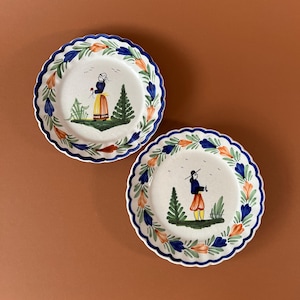 Könnte beinhalten: Zwei runde Keramikteller mit gewelltem Rand, verziert mit einer handbemalten Bordüre aus blauen und orangefarbenen Blumenmotiven. In der Mitte jedes Tellers ist eine Figur in traditioneller Kleidung in einer Landschaft mit grünen Bäumen abgebildet.
