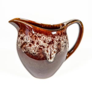 Puede incluir: Una jarra de cerámica marrón con asa y pico vertedor. La jarra tiene un acabado brillante con un diseño moteado blanco y marrón en la mitad superior y una base marrón sólida. El asa es curva y el pico es puntiagudo.