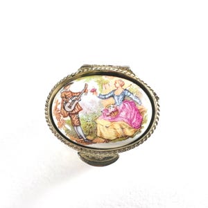 May include: Oval trinket box with a detailed scene of a man playing a guitar and a woman in a pink dress. The box has a gold-colored metal frame with a rope-like border and a small base.