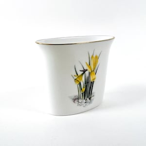 May include: White ceramic vase with a gold rim and a floral design of yellow daffodils.