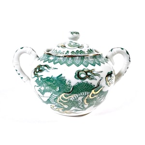 Op de afbeelding: Witte keramische suikerpot met een deksel en twee handvatten. De kom is versierd met een groen drakenontwerp en gouden accenten. Het deksel heeft een bijpassend groen en goud patroon.