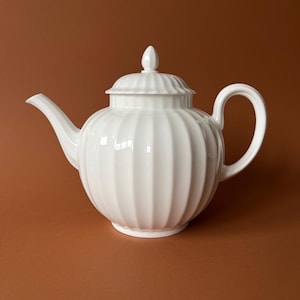 May include: A white ceramic teapot featuring a rounded, bulbous body with vertical fluted ribbing. The teapot includes a matching lid with a pointed finial, a curved handle on the right side, and a tapered spout extending from the left side.