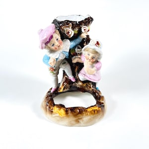 Może przedstawiać: Figurka z porcelany w stylu vintage przedstawiająca chłopca i dziewczynkę wokół pnia drzewa. Chłopiec ma na sobie niebiesko-biały strój z różowym kapeluszem, a dziewczynka różową sukienkę i biały kapelusz. Podstawa jest brązowa, a drzewo ma róże.