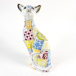 May include: A ceramic cat figurine with a patchwork design. The cat is white with a colorful patchwork pattern of squares, hearts, and swirls. The ears have gold trim. The cat is sitting upright with its head tilted slightly upward.