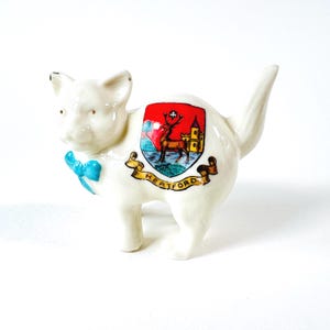 Puede incluir: Figura de gato de cerámica blanca con pajarita azul. El gato tiene un emblema colorido en su costado con un ciervo, un castillo y la palabra "Hertford". La figura está sobre una superficie blanca.