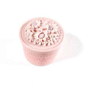 Op de afbeelding: Een kleine, ronde, roze sieradendoos met een bloemmotief op het deksel. Het deksel heeft een gedetailleerde opstelling van gebeeldhouwde bloemen. De doos heeft een textuurpatroon rond de zijkanten. Het algehele ontwerp is delicaat en decoratief.