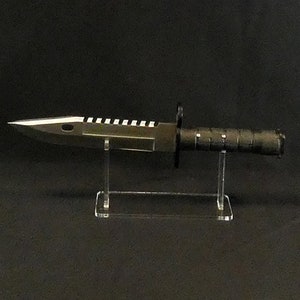 Könnte beinhalten: Ein schwarzes und silbernes Messer mit einer gezahnten Klinge wird auf einem klaren Acrylständer präsentiert. Das Messer hat einen schwarzen Griff mit einer strukturierten Grifffläche.