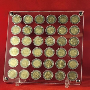 May include: A clear acrylic display case holding a collection of Euro coins. The coins are gold and silver in color, each featuring unique designs and symbols. The case is square and has a clear stand, showcasing the coins against a red background. A numismatic collectible.
