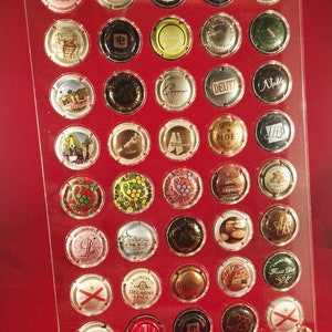 Puede incluir: Un expositor de acrílico transparente con 36 compartimentos circulares, cada uno con un tapón de botella de champán diferente. Los tapones presentan varios logotipos y diseños, incluidos los nombres "Moët Chandon", "Deutz", "Champagne Delagne & Fils" y "Ruinart".