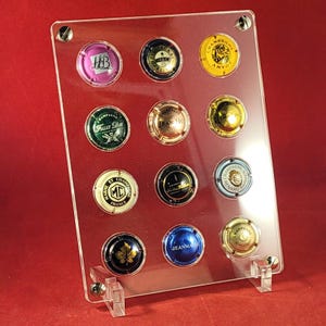 Puede incluir: Una vitrina de acrílico transparente con 12 tapones de botellas de champán. Los tapones están dispuestos en forma de cuadrícula y presentan varios diseños y colores, incluyendo dorado, plateado y rojo.