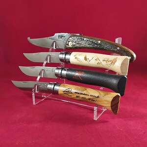 K&ouml;nnte beinhalten: Vier verschiedene Taschenmesser mit Holzgriffen, die auf einem durchsichtigen Acrylst&auml;nder pr&auml;sentiert werden. Die Messer haben unterschiedliche Designs und Farben, darunter ein schwarzer Griff mit wei&szlig;em Logo, ein Holzgriff mit einer Bergszene und ein Holzgriff mit einem braunen und schwarzen Muster.
