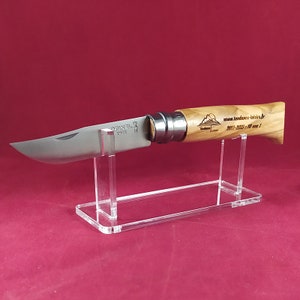 Op de afbeelding: Een vouwmes met een houten handvat en een roestvrijstalen mes, gegraveerd met de merknaam "Opinel" en de tekst "www.tendances-latins.fr 2011-2021 - 10 ans 1". Het mes wordt gepresenteerd op een heldere acrylstandaard.