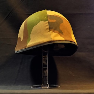 Peut inclure: Un casque de camouflage avec une bordure noire, présenté sur un support en acrylique transparent.
