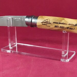Op de afbeelding: Een transparante acryl messenstandaard met een Opinel mes met een houten handvat. Het mes heeft een roestvrijstalen lemmet en het handvat is gegraveerd met de tekst "Tendance Loisirs 2011-2021: 10 ans!" en een logo.