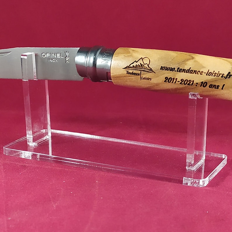 Op de afbeelding: Een transparante acryl messenstandaard met een Opinel mes met een houten handvat. Het mes heeft een roestvrijstalen lemmet en het handvat is gegraveerd met de tekst "Tendance Loisirs 2011-2021: 10 ans!" en een logo.
