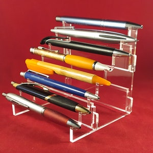 May include: A clear acrylic pen holder with seven pens of various colours and styles. The holder has six tiers, each with a slot for a pen. The pens are arranged in a staggered pattern.