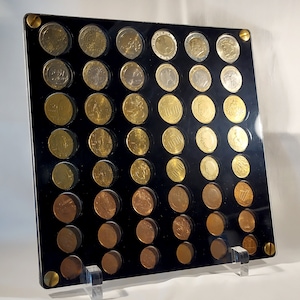 Puede incluir: Una vitrina para monedas de acrílico negro con un frente de acrílico transparente. La vitrina contiene 48 monedas de euro dispuestas en una cuadrícula. Las monedas están dispuestas en filas y columnas, con cada moneda en su propio compartimento individual. La vitrina tiene un soporte de acrílico transparente que permite que se exhiba en una mesa o estante.