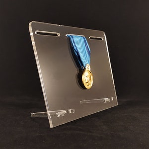 Peut inclure: Un présentoir en acrylique transparent tenant une médaille dorée avec un ruban bleu. La médaille présente un portrait et est suspendue en haut du présentoir. Le présentoir est incliné et a un design simple et moderne.
