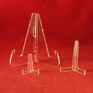 May include: Clear acrylic display stands in different sizes. The stands have a triangular or T-shaped base and a hinged support for holding items. The stands are designed to showcase plates, artwork, or other collectibles. The background is a solid red.