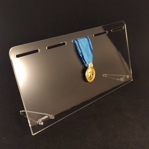 Pode incluir: Um suporte de exibição de medalhas de acrílico transparente com cinco ranhuras para pendurar medalhas. Uma medalha de ouro com uma fita azul está pendurada em uma das ranhuras.