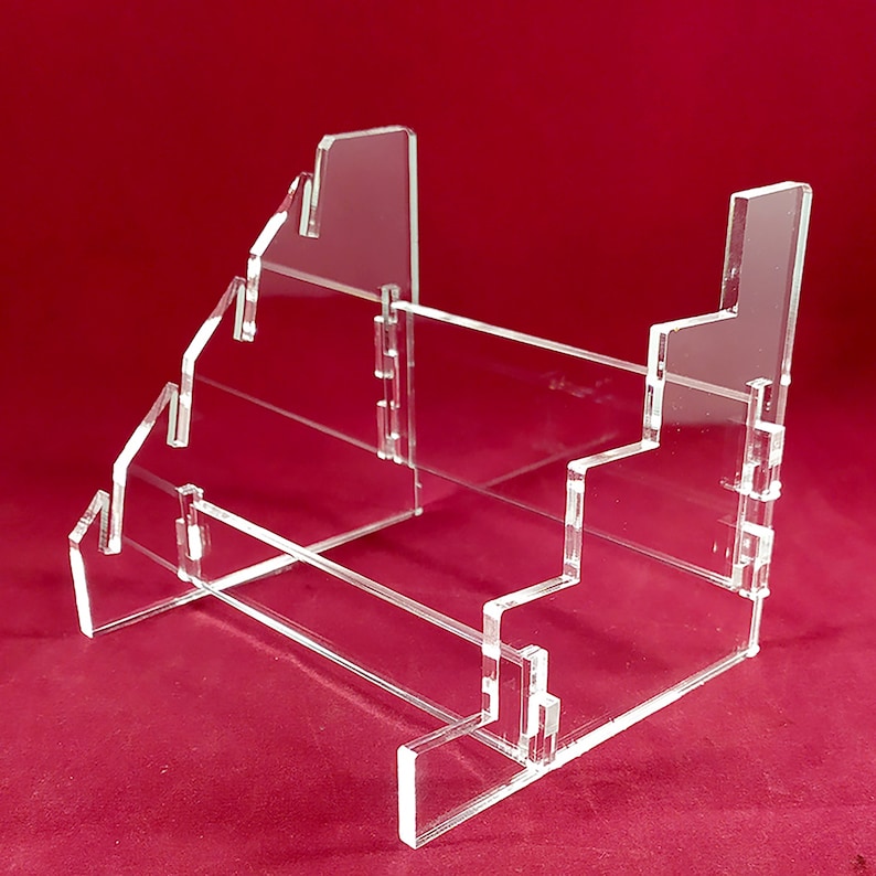 K&ouml;nnte beinhalten: Ein transparenter Acryl-Pr&auml;sentationsst&auml;nder mit drei Ebenen zum Ausstellen von Gegenst&auml;nden wie B&uuml;chern, Karten oder anderen Sammlerst&uuml;cken. Der St&auml;nder ist aus klarem Acryl gefertigt und hat ein modernes, minimalistisches Design.