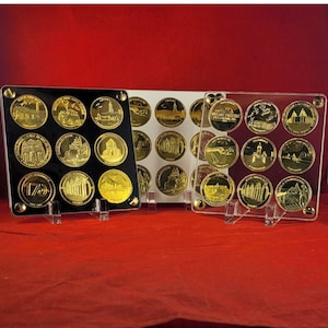 Op de afbeelding: Drie sets goudkleurige herdenkingsmunten tentoongesteld in transparante acrylhouders. Elke munt heeft een gedetailleerd reliëfontwerp. De sets zijn gerangschikt op een rode ondergrond, met een donkere achtergrond.