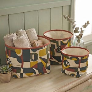 Peut inclure: Trois paniers de rangement cylindriques en tissu avec un motif géométrique jaune moutarde, rouge foncé, bleu marine et crème. Les paniers présentent une bordure unie de couleur rouille sur le dessus et une doublure intérieure crème.