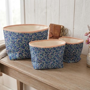 Peut inclure: Un ensemble de trois paniers de rangement en tissu de tailles variées, présentant un extérieur bleu foncé avec un motif floral dense dans des tons de rose, jaune et vert, fini avec un bord rose clair uni et une doublure intérieure blanc cassé.