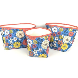 Puede incluir: Tres cubos de almacenamiento de tela de diferentes tamaños. Presentan un estampado floral azul con flores blancas, amarillas y coral. Los bordes son de color coral y los interiores son blancos. Estos cubos son ideales para organizar objetos.